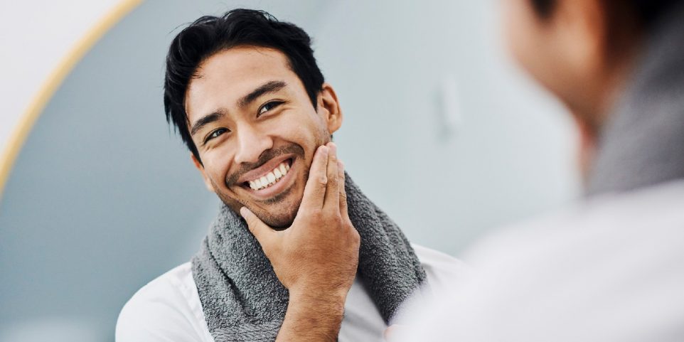 Man happy about face skin care product for healthy body and facial skincare for guys. Handsome young male looking in the mirror with toothy smile, touching his beard satisfied with his shaving cream