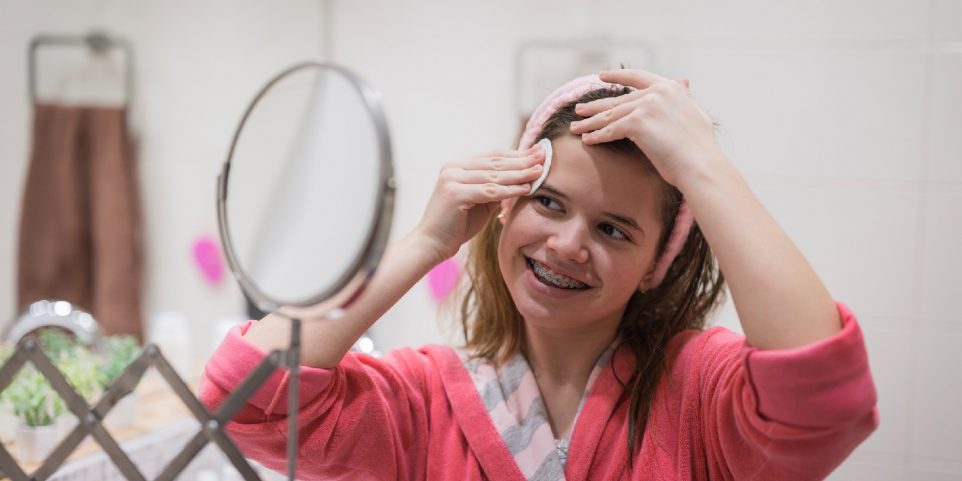 Cheerful young girl cleaning forehead with cotton pad and micellar water