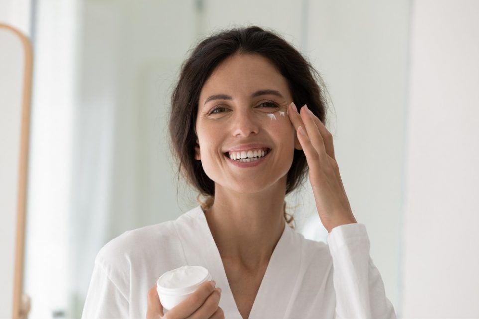 Head shot portrait happy Hispanic woman in white bathrobe smile looks at camera holds jar applying cream on cheek area.