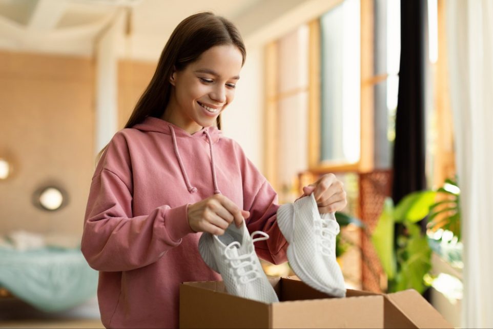 Happy teen girl holding new footwear unpacking cardboard box, receiving shoes as a gift