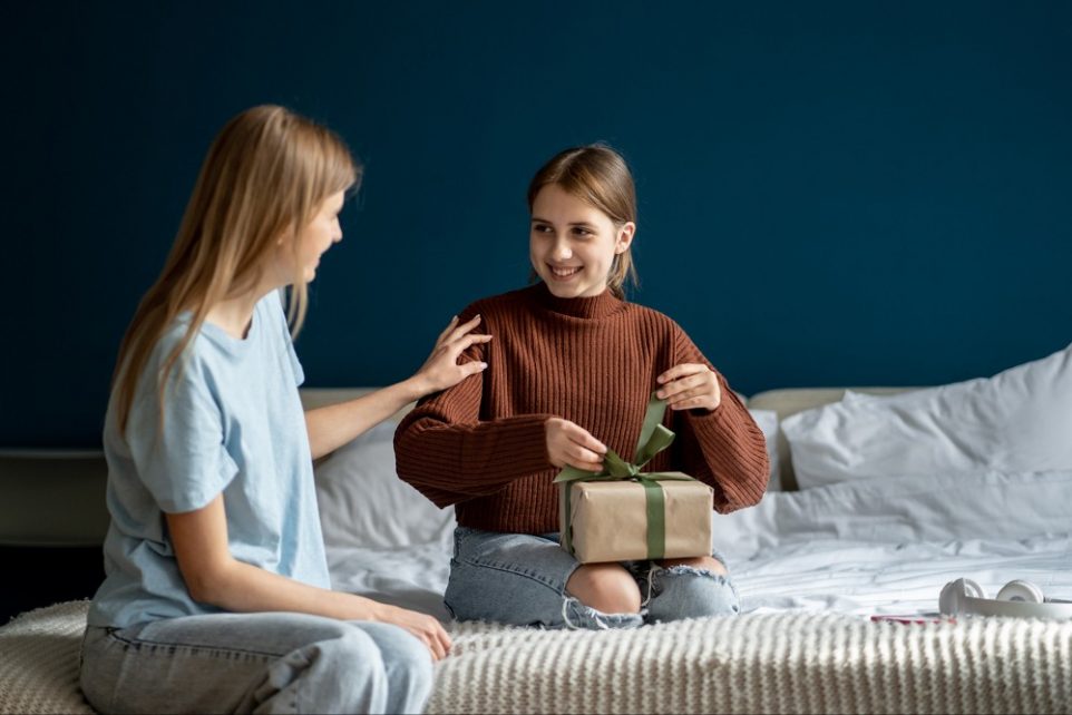 Pleased smiling teenage girl interested untie bow on box birthday gift from caring happy mom at home